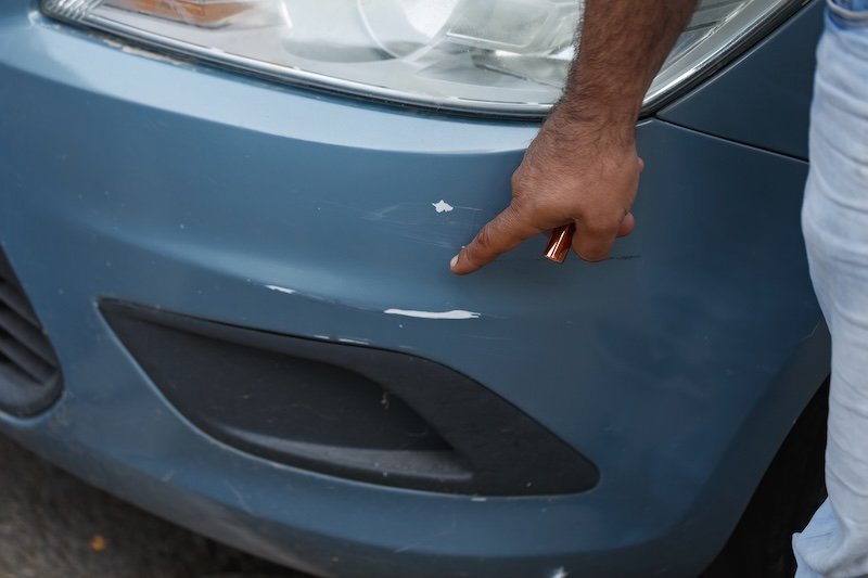 A person man is pointing at a scratch and small dent on the front bumper of a blue car. The focus is on the damaged area near the headlight, highlighting the chipped paint and surface scuff marks.