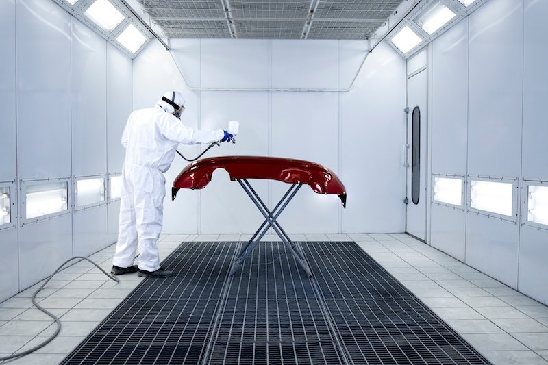 Car painter in protective clothes and mask painting automobile bumper with metallic paint and varnish in chamber workshop.